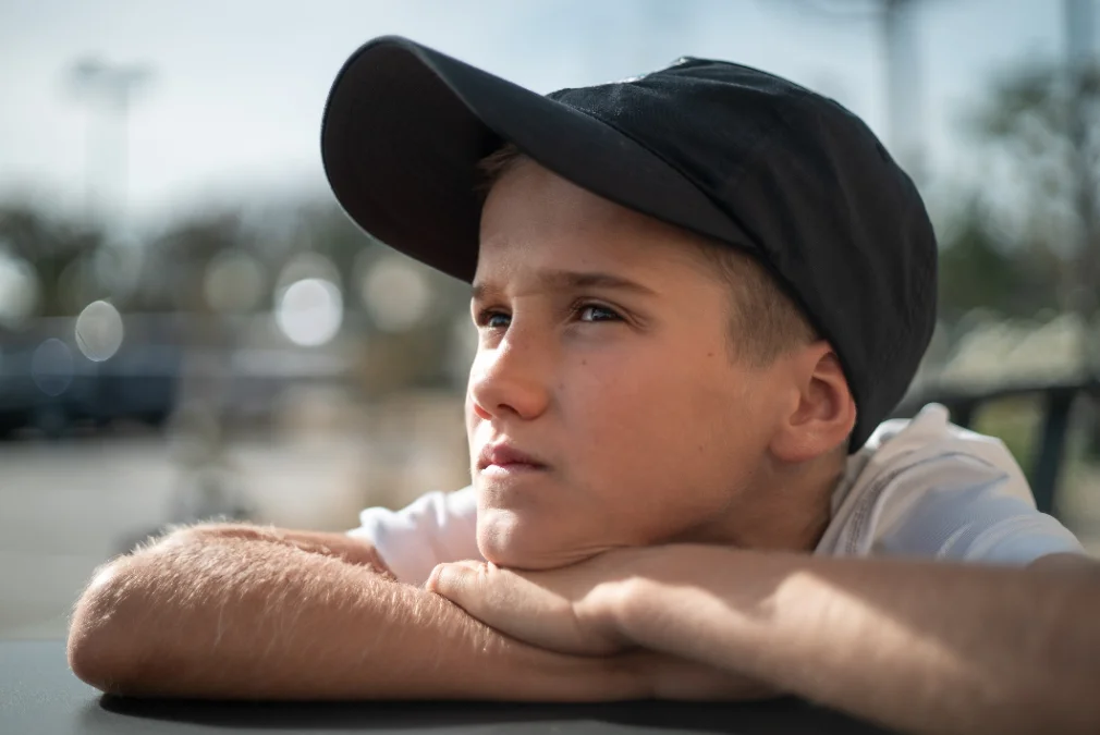 A boy in a baseball cap rests his chin on his arms, gazing into the distance.