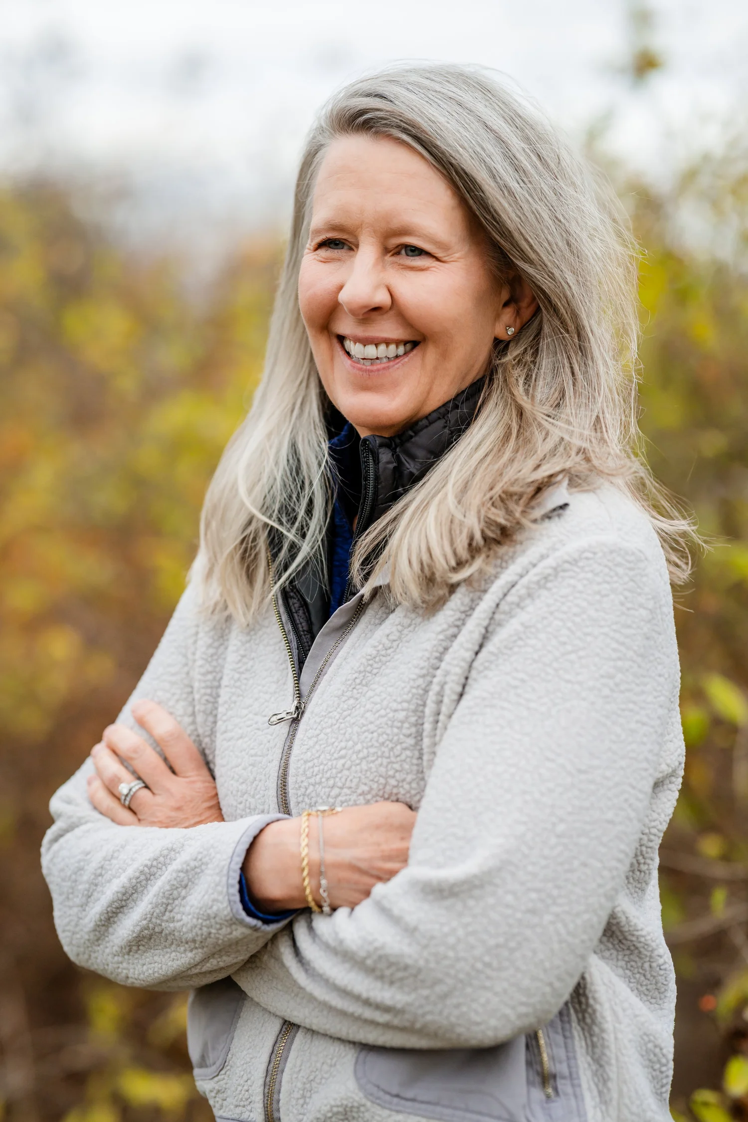 Clara Bowron smiling with her arms crossed, standing outdoors in front of autumn brush.