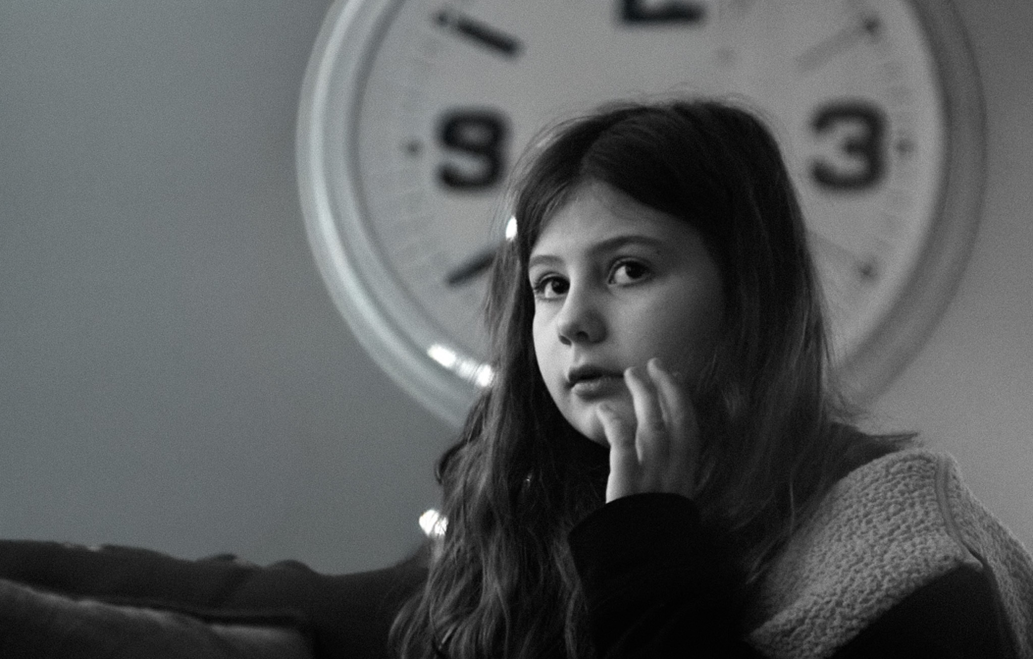 A girl rests her chin on her hand, looking away, with a large wall clock behind her.